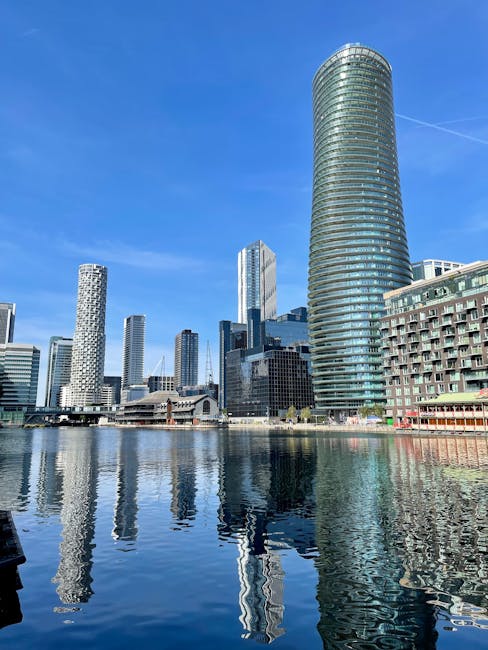 A view of a modern cityscape along a waterfront featuring high-rise office buildings with glass facades reflecting the sky and surroundings. In the foreground, a dockside area with a white metal gangw